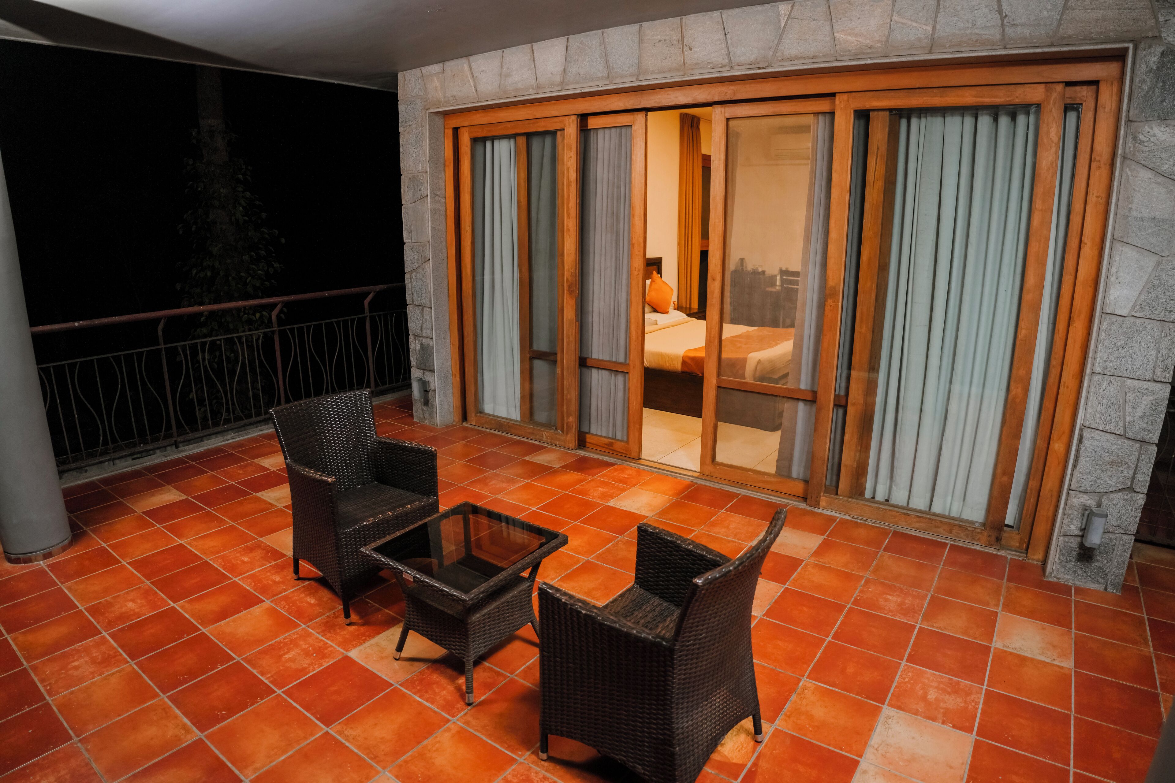 A balcony with wicker furniture, a glass table, and a view of a room with a bed and curtains.