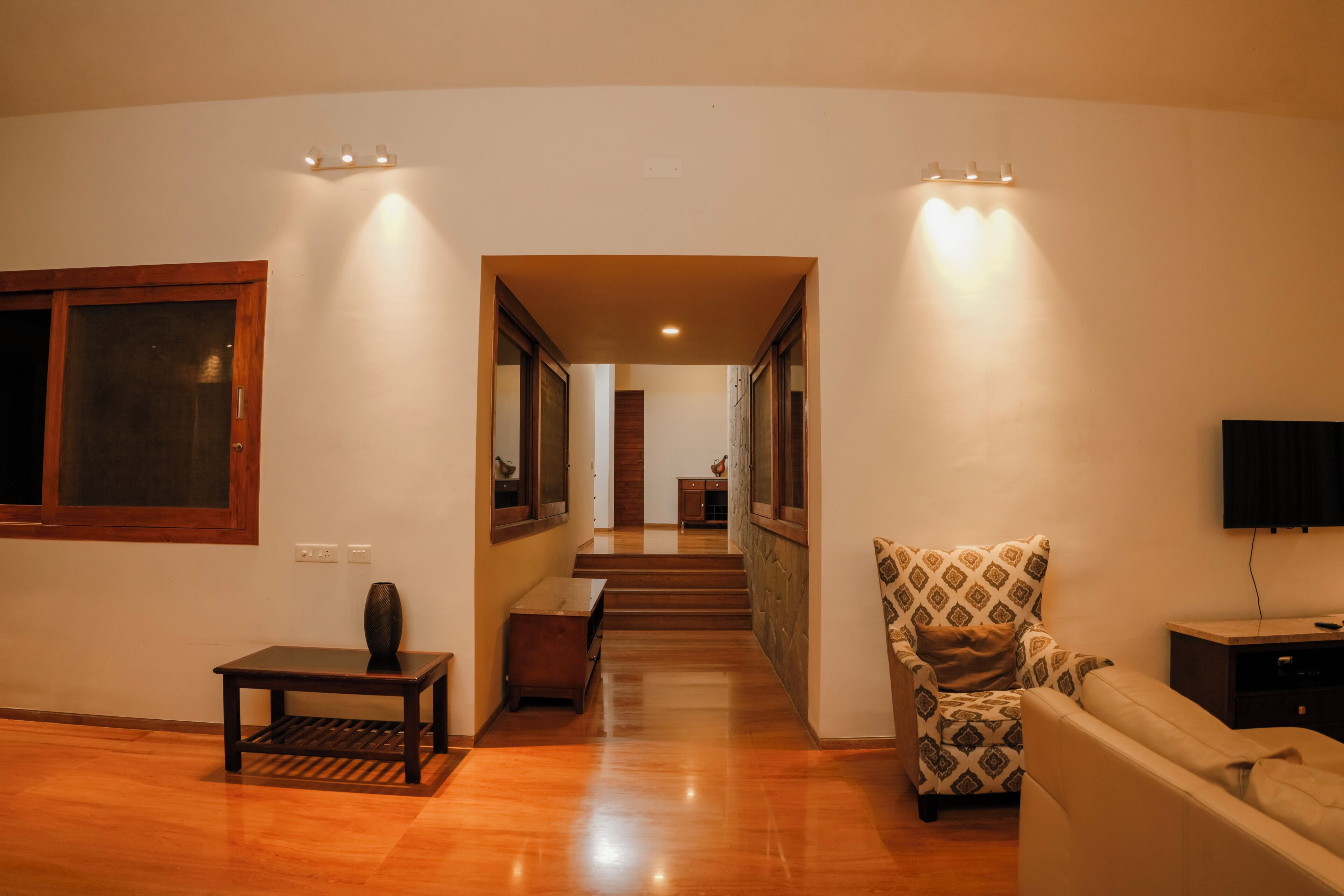 A spacious living area with a wooden floor, a sofa, a patterned armchair, a small table, and a television.