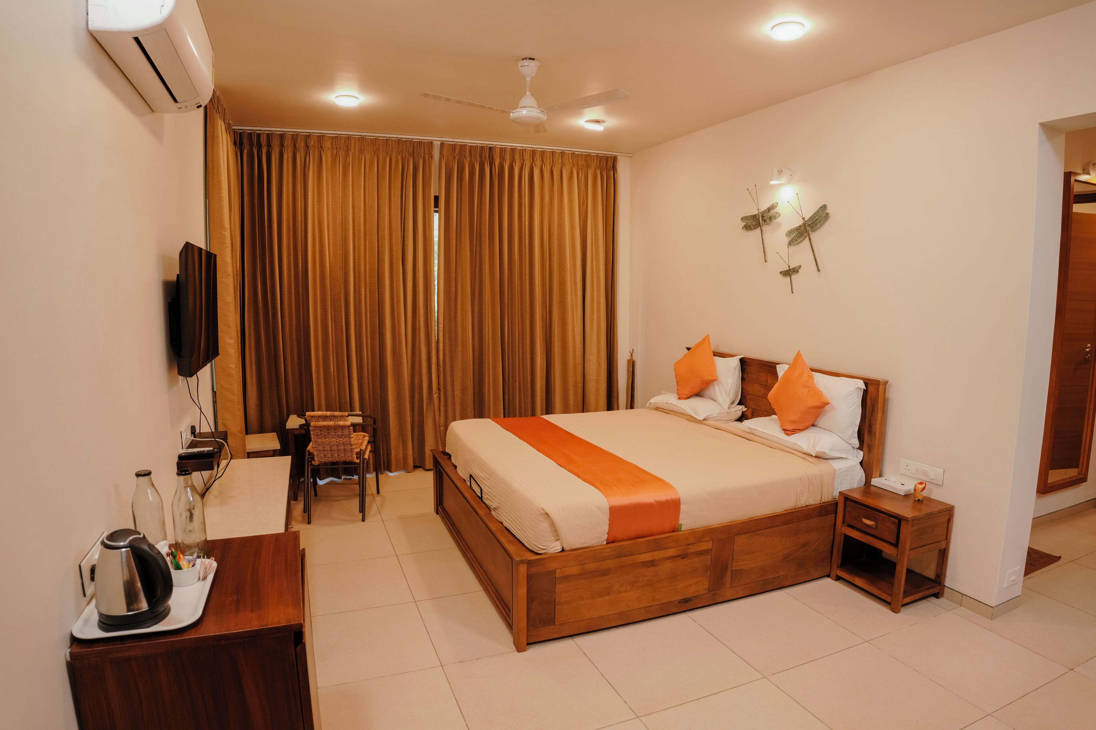 A hotel room with a wooden bed, a desk with a kettle, a chair, and a TV mounted on the wall.