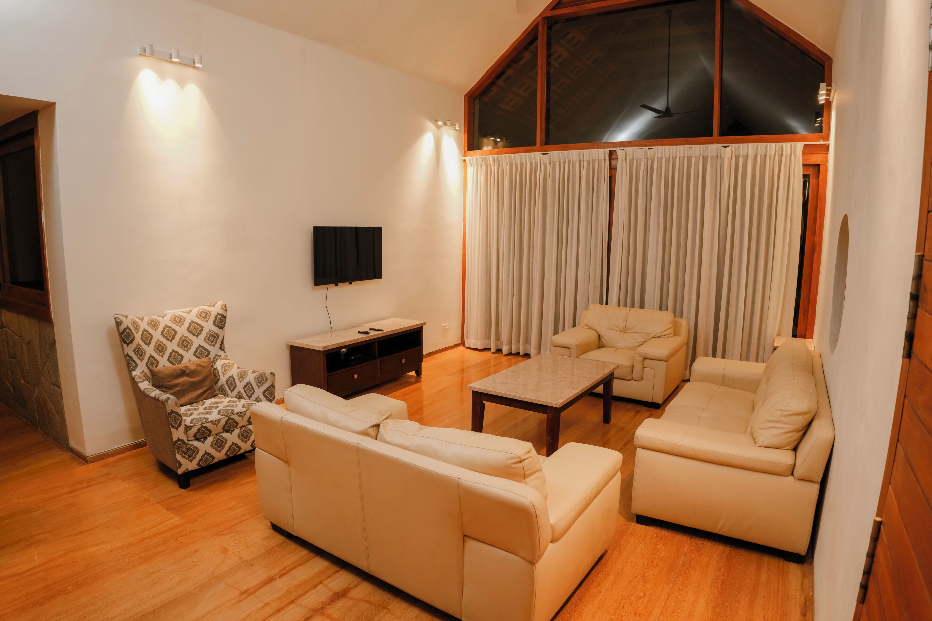 Sala de estar com sofá, poltrona e mesa de centro, uma TV fixada na parede e uma janela grande com cortinas.