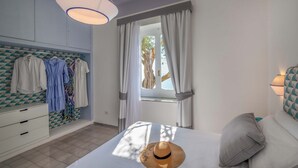 A bedroom with a bed, a window with curtains, a wardrobe with hanging clothes, and a ceiling light. - Villa Pelagium on the Amalfi Coast (Praiano)