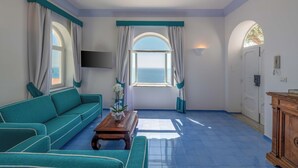 A room with a teal sofa, a wooden coffee table, a TV mounted on the wall, and a view of the sea through a window with teal curtains. - Villa Pelagium on the Amalfi Coast (Praiano)
