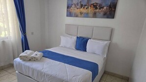 En snyggt bäddad säng med vita och blå sängkläder, en målning med en stadsbild på väggen och ett fönster med blå gardiner.