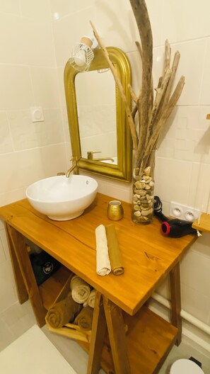 Un baño con tocador de madera, un lavabo blanco, un espejo con marco dorado y un florero con ramas.