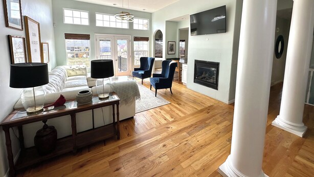 A spacious living room with a white sofa, a blue armchair, and a wooden coffee table. There is a fireplace and a large window with a view of the outdoors.