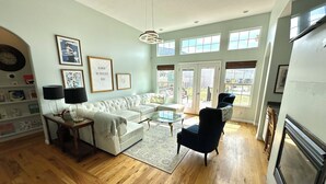 A spacious living room with a white sofa, a glass coffee table, and a blue armchair. There are framed pictures on the walls and a chandelier hanging from the ceiling.