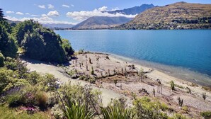 A lakeside view with a clear sky, mountains in the background, and a sandy beach area with sparse vegetation. - Amazing Lakeside Apartment (Queenstown)