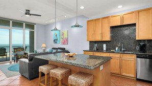 A modern kitchen with granite countertops, wooden cabinets, and a large island with stools. There is a dining area with a view of the ocean. - 3rd Coast Getaways - Sapphire 803 (South Padre Island)