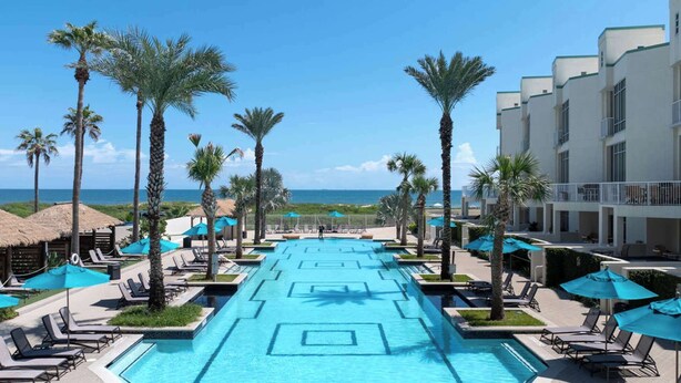 A resort pool with clear blue water, surrounded by palm trees and lounge chairs, overlooking the ocean. - 3rd Coast Getaways - Sapphire 803 (South Padre Island)
