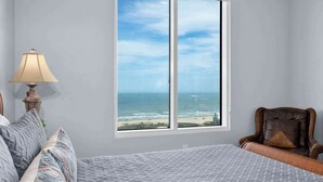 A bedroom with a large window overlooking the ocean, a bed with patterned bedding, a chair, and a lamp. - 3rd Coast Getaways - Sapphire 803 (South Padre Island)