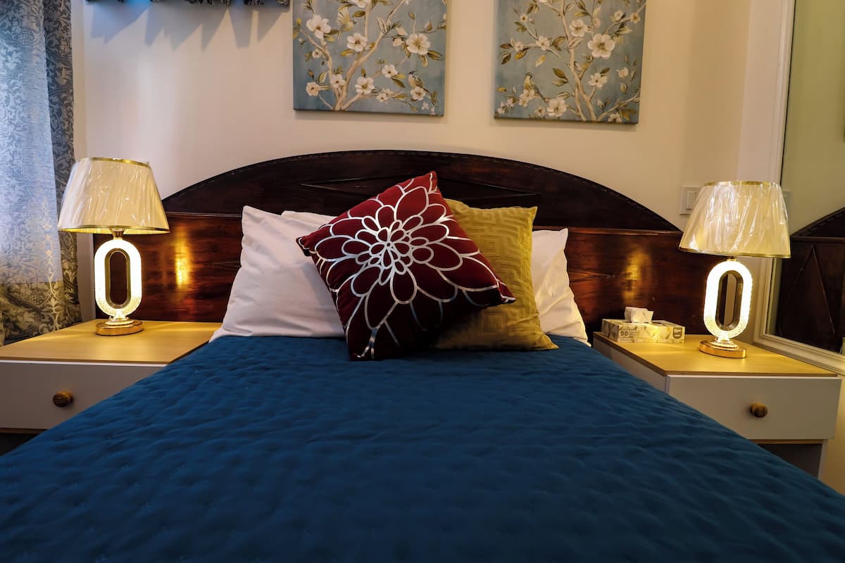 A neatly made bed with a blue bedspread, a red floral pillow, and two bedside lamps on wooden nightstands.