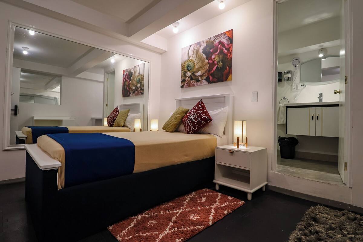 A modern bedroom with a large bed, bedside tables, a mirror, and a bathroom visible through an open door.
