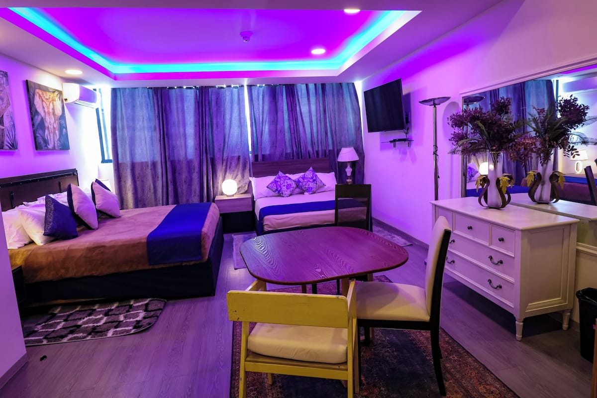 A modern hotel room with a bed, bedside table, lamp, and a small dining table with chairs.