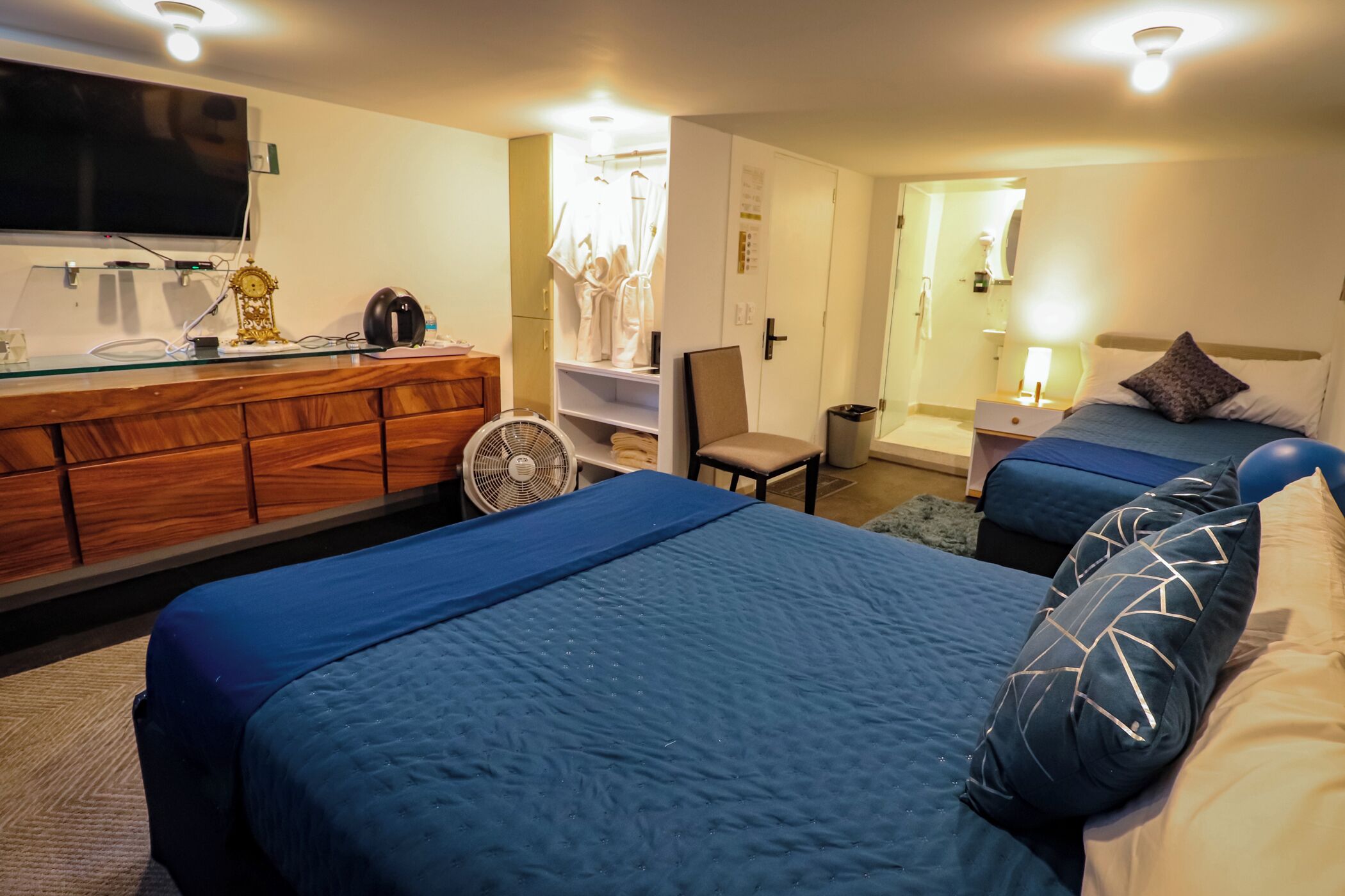 A hotel room with two beds, a TV mounted on the wall, a desk with a fan, and a wardrobe.