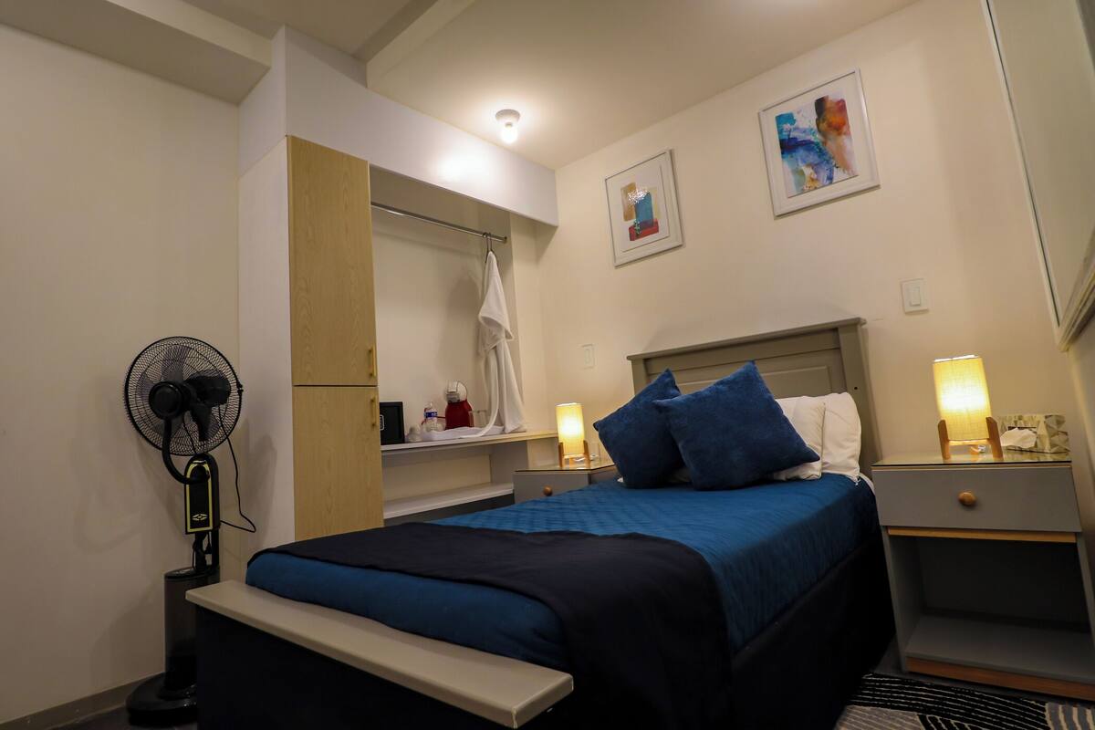 A hotel room with a bed, bedside tables, a fan, a bench, a mirror, and artwork on the walls.