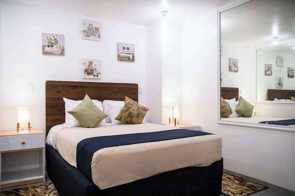 A neatly arranged bedroom with a wooden headboard, a bed with a beige and navy blue bedspread, and two bedside lamps. There are three framed artworks on the wall above the bed.