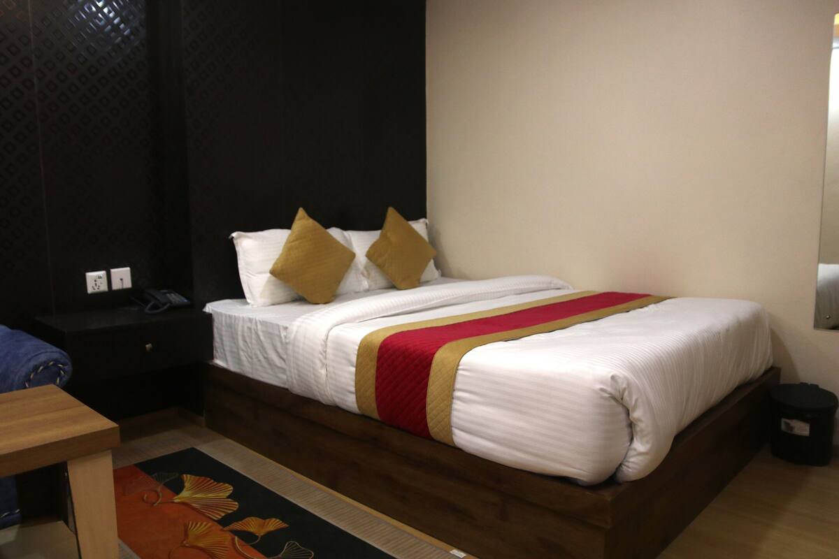 A neatly made bed with white bedding and a red and gold striped blanket, a wooden nightstand with a lamp, and a patterned rug.
