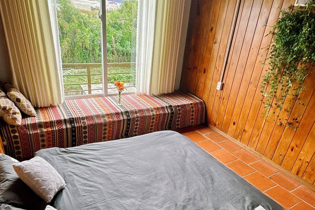 A bedroom with a large bed, a window with curtains, a wooden wall, and a hanging plant.