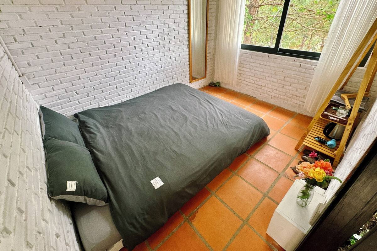 A small, compact room with a brick wall, a single bed with grey bedding, a small table with a vase of flowers, and a window with a view of trees.