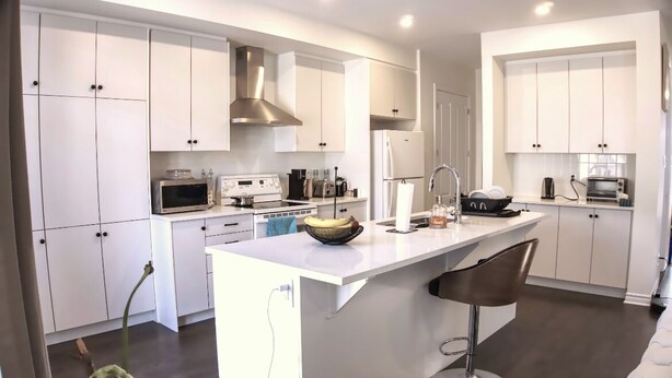 A modern kitchen with white cabinetry, a central island, and bar stools. - Spacious and Cozy Room with Private Bathroom in a house with WiFi, AC in Ottawa. (Ottawa)