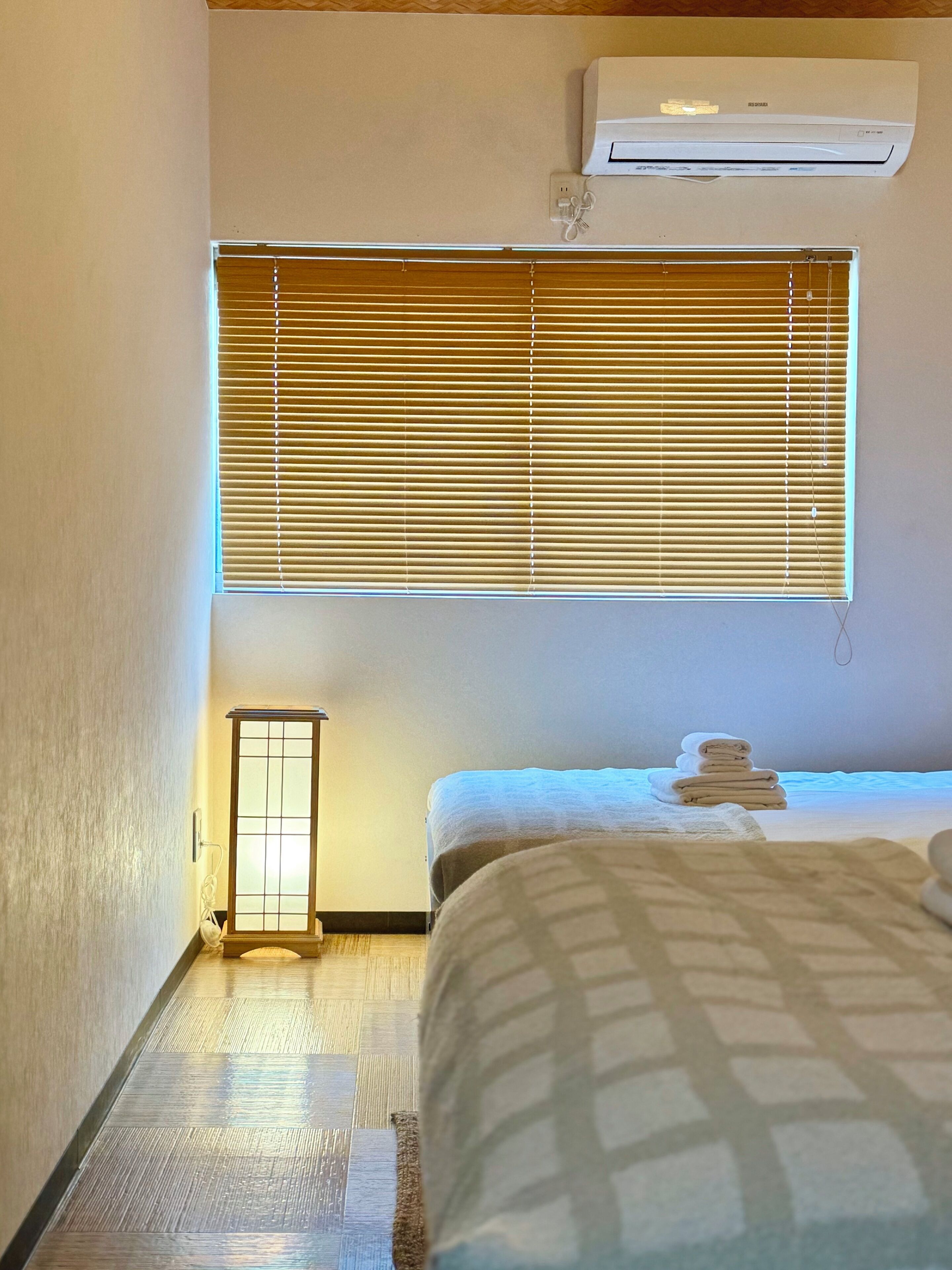 A hotel room with a bed, a window with blinds, a wall-mounted air conditioner, and a small table with a lamp.