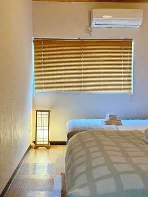 A hotel room with a bed, a window with blinds, a wall-mounted air conditioner, and a small table with a lamp. - Miyu Kitagatsukyo (Osaka)