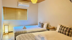 Een hotelkamer met twee bedden, een raam met zonwering, een airconditioner, een nachtkastje met een lamp, en een receptie.