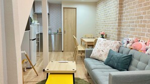 Een gezellige woonkamer met een grijze bank, een gele salontafel en een wand met een bakstenen patroon. Op de achtergrond is een keuken zichtbaar met een witte koelkast en een houten deur.