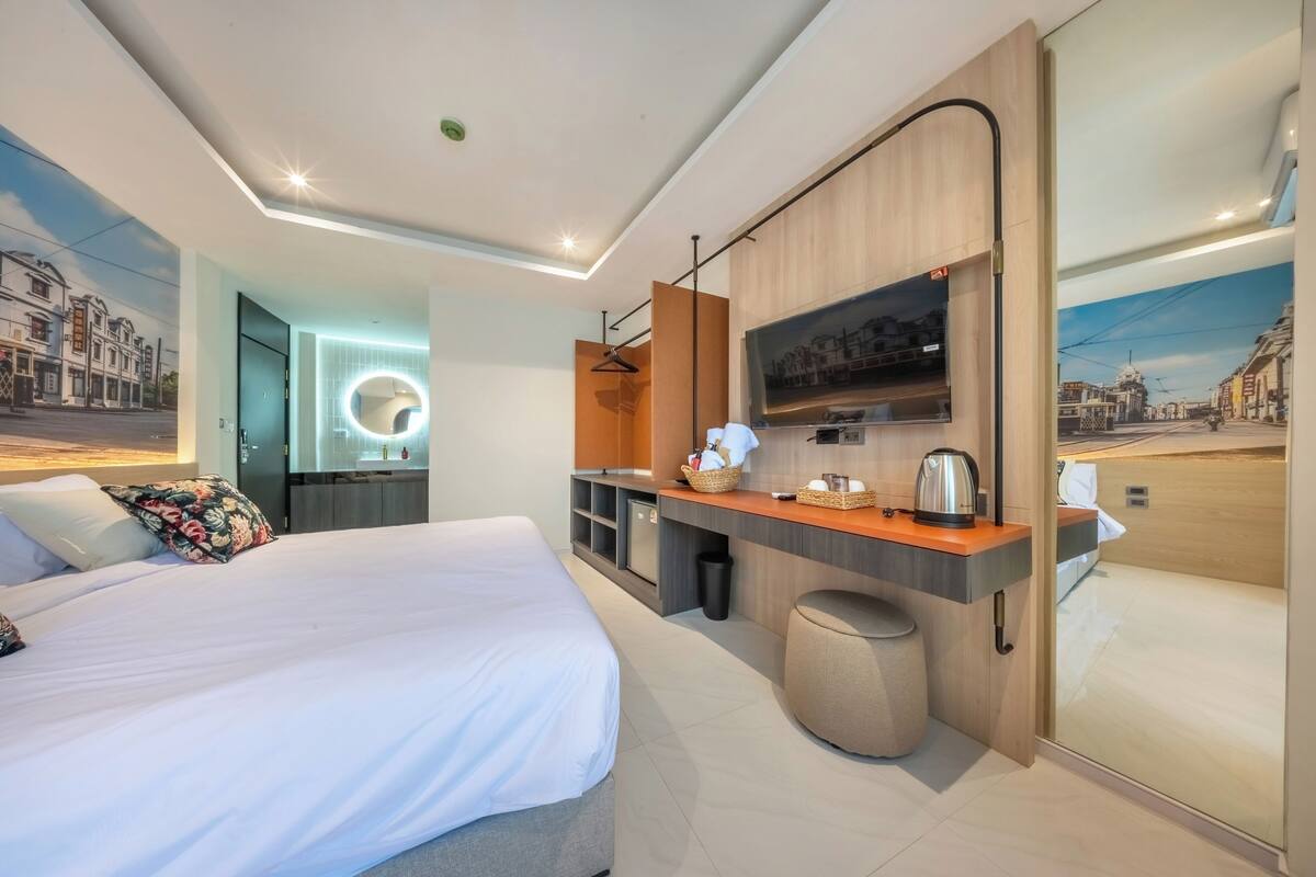 A modern hotel room with a large bed, a small kitchenette, a TV, and a view of a cityscape.