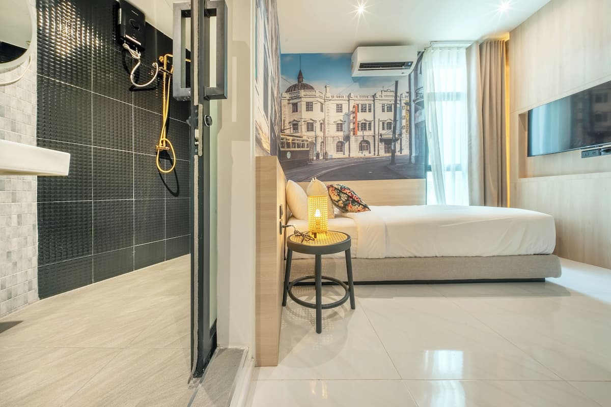 A modern hotel room with a bed, a small table with a lamp, a shower area, and a mural of a cityscape on the wall.
