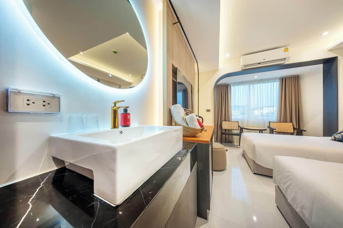 A modern hotel room with a large white sink, two beds, a seating area with a table and chairs, and a large mirror.