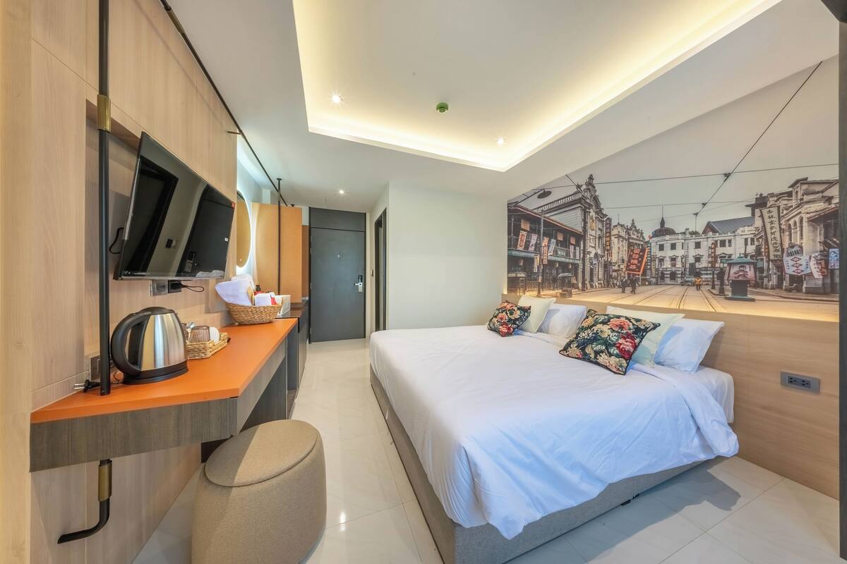 A modern hotel room with a bed, a desk with a kettle, a flat-screen TV, and a mural of a city street.