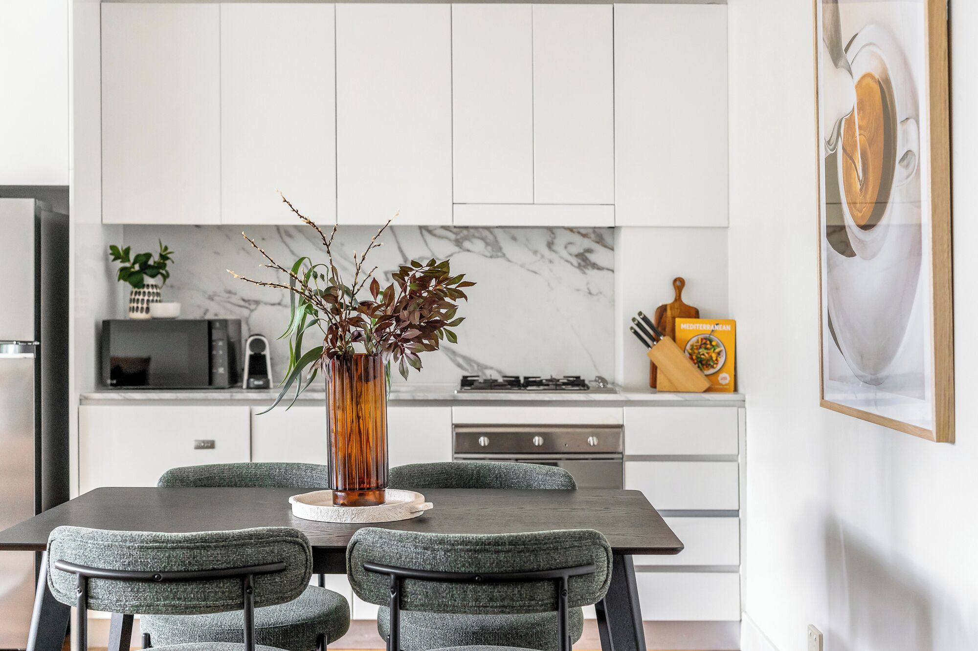 A modern kitchen with white cabinets, a marble backsplash, a dining table with four chairs, and a vase with branches on the table.