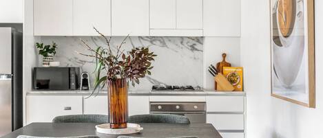 A modern kitchen with white cabinets, a marble backsplash, a dining table with four chairs, and a vase with branches on the table.