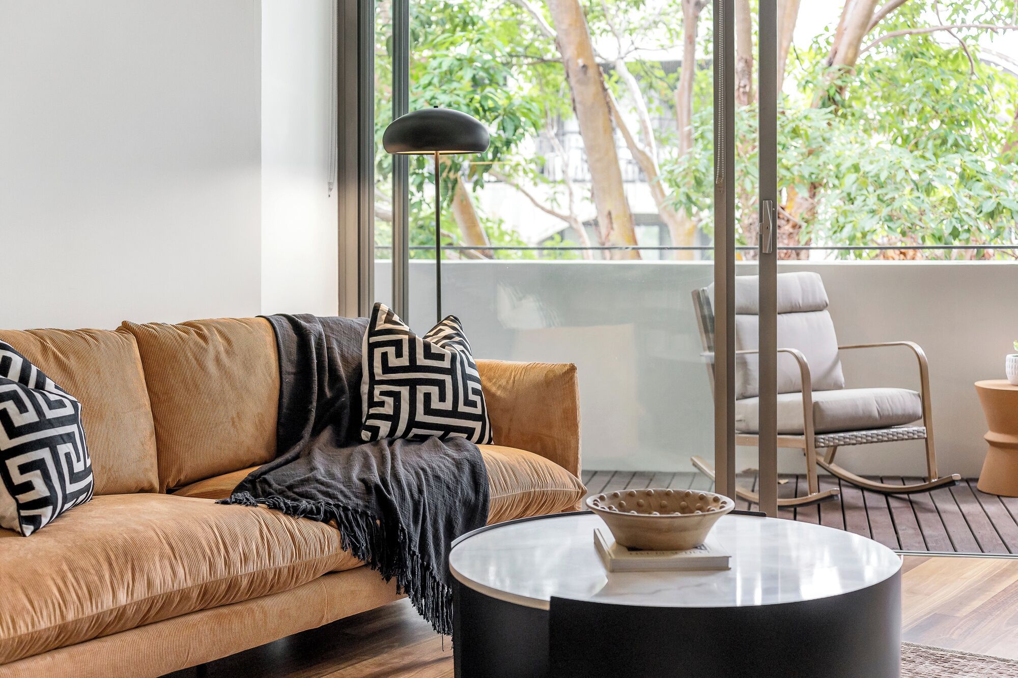 A living room with a sofa, a coffee table, a floor lamp, and a view of greenery outside.