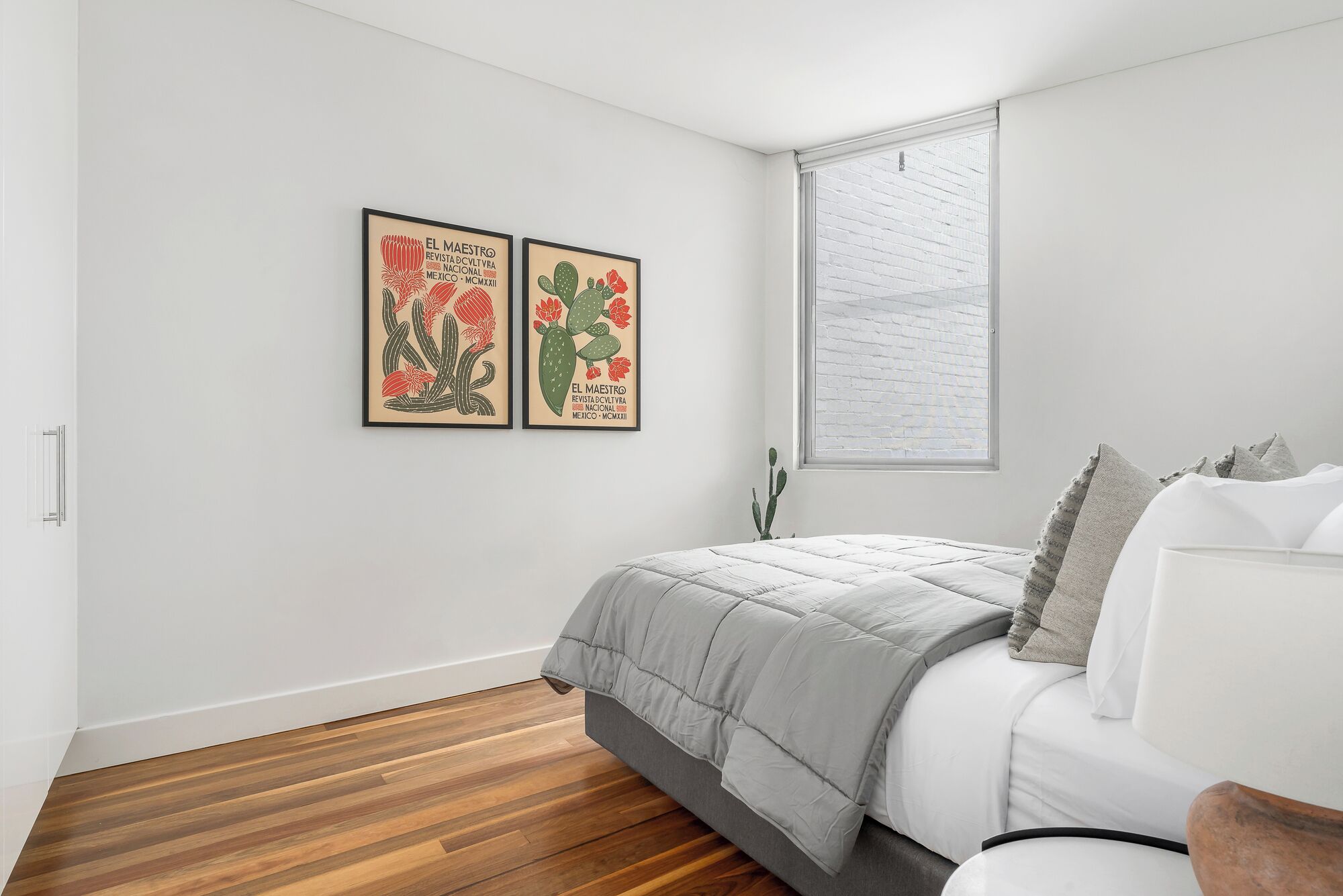 A bedroom with a bed, wooden flooring, a window with blinds, and two framed artworks on the wall.
