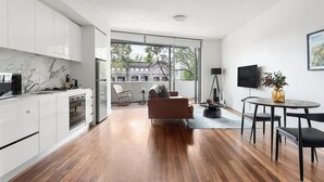A modern kitchen with white cabinets and a marble backsplash, a dining area with a round table and chairs, and a large window offering a view of a building and trees.