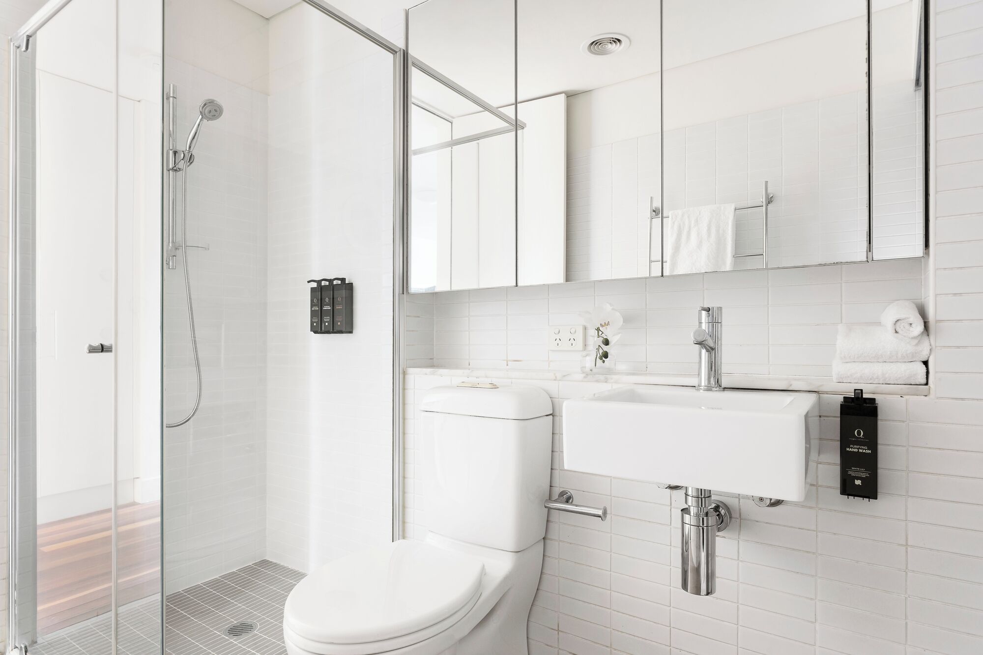 A modern bathroom with a glass shower enclosure, a white toilet, a sink with a hand wash station, and a mirror above the sink.