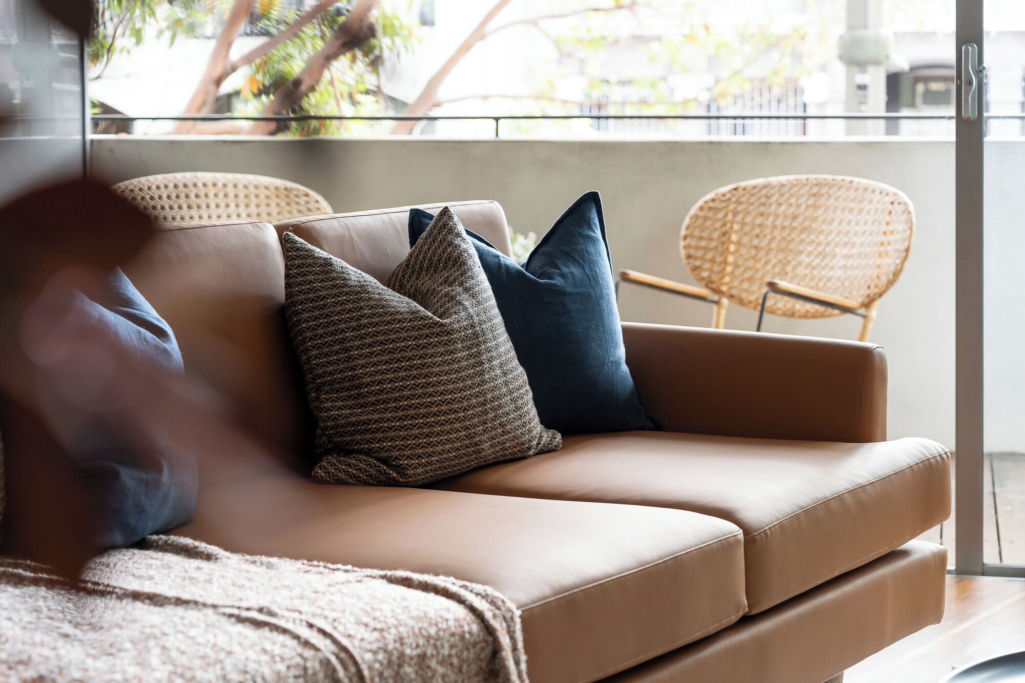 A tan leather sofa with two pillows, a woven throw blanket, and a wicker chair in a bright room with a view of trees and buildings.
