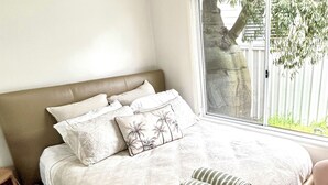 A bedroom with a large bed, multiple pillows, a window with a view of a tree and fence, and a wooden floor. - 21A Boab Cottage (Mayfield)