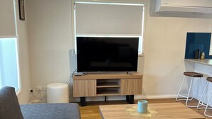 A modern living room with a flat-screen TV, wooden coffee table, grey sofa, and a small bar area with stools. - 21A Boab Cottage (Mayfield)