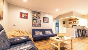 Una sala de estar con un sofá azul oscuro, un sillón de cuero y una mesa de centro de madera. Se observan cuadros enmarcados en la pared y una zona de cocina al fondo.