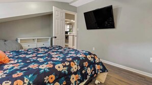Un dormitorio con una cama, un televisor en la pared y una puerta que da a una zona de cocina.
