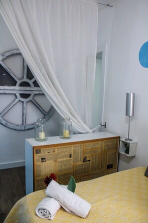 Una cama bien hecha con una colcha amarilla estampada, una toalla blanca y una flor decorativa. Hay un aparador de madera con un espejo arriba, dos frascos de vidrio con velas y una cortina blanca.