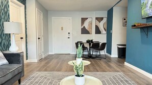 Un salon moderne avec un canapé gris, une table basse ronde agrémentée d’une plante, un coin repas avec une table et des chaises, et des œuvres d’art abstraites accrochées au mur.