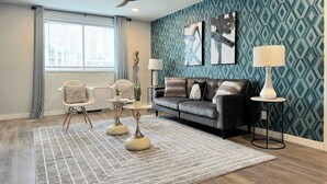 Un salon moderne avec un canapé gris, deux fauteuils, un tapis à motifs, un ventilateur de plafond et des œuvres d’art accrochées au mur.