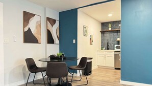 Un espace de repas moderne avec une table ronde et quatre chaises, une cuisine avec des meubles blancs et un mur d’accent bleu, et des œuvres d’art abstraites accrochées aux murs.