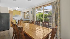 Un coin repas avec une table et des chaises en bois, une cuisine à l’arrière-plan et une vue sur la verdure à l’extérieur.