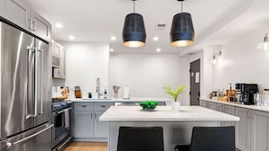 A modern kitchen with stainless steel appliances, a central island with two chairs, pendant lighting, and a small plant. - Sleeps 12, Mxn Streets (Pittsburgh)