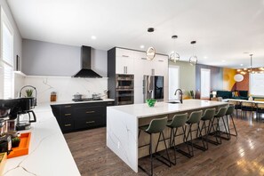 A modern kitchen with a central island, bar stools, and a dining area with a colorful wall. - ★ HostWise Stays ★ Stylish Homes with Rooftop Decks, BBQ & Parking (Pittsburgh)